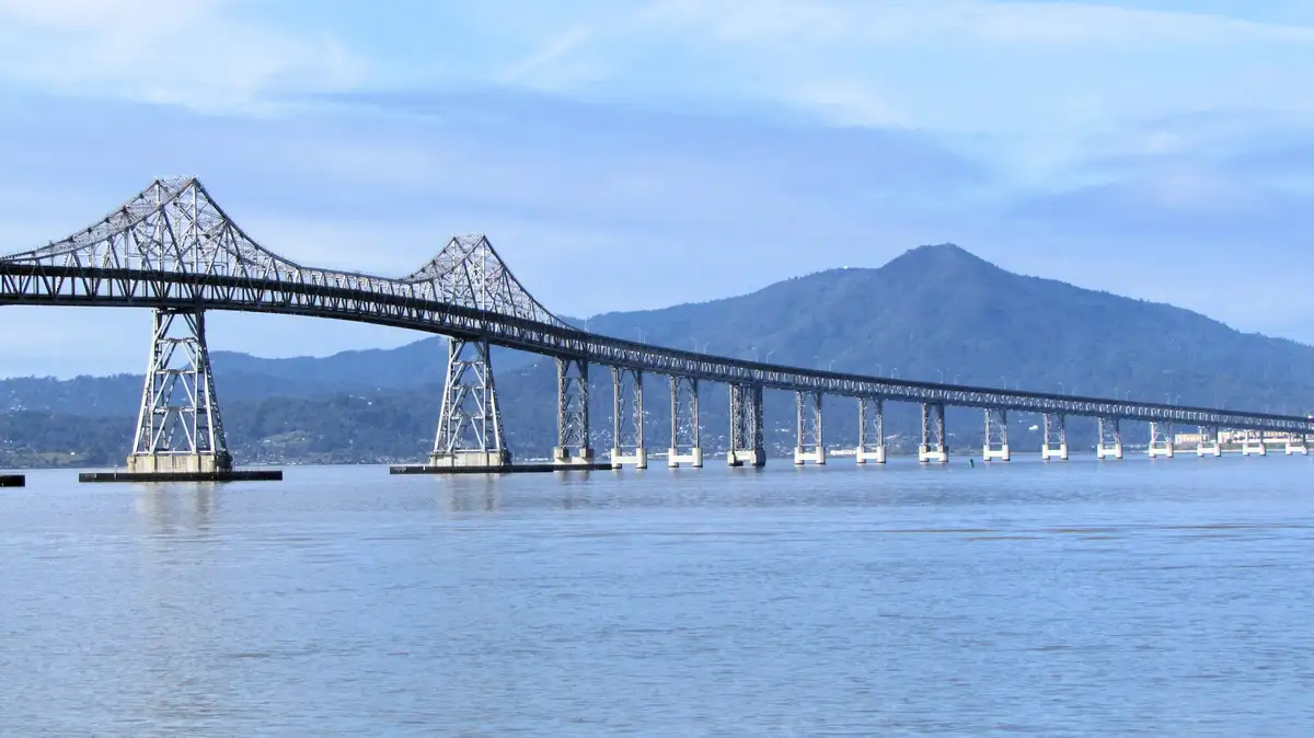 A wide-angle photograph shows a long, steel truss bridge spanning a calm blue body of water, with distant green hills and a…