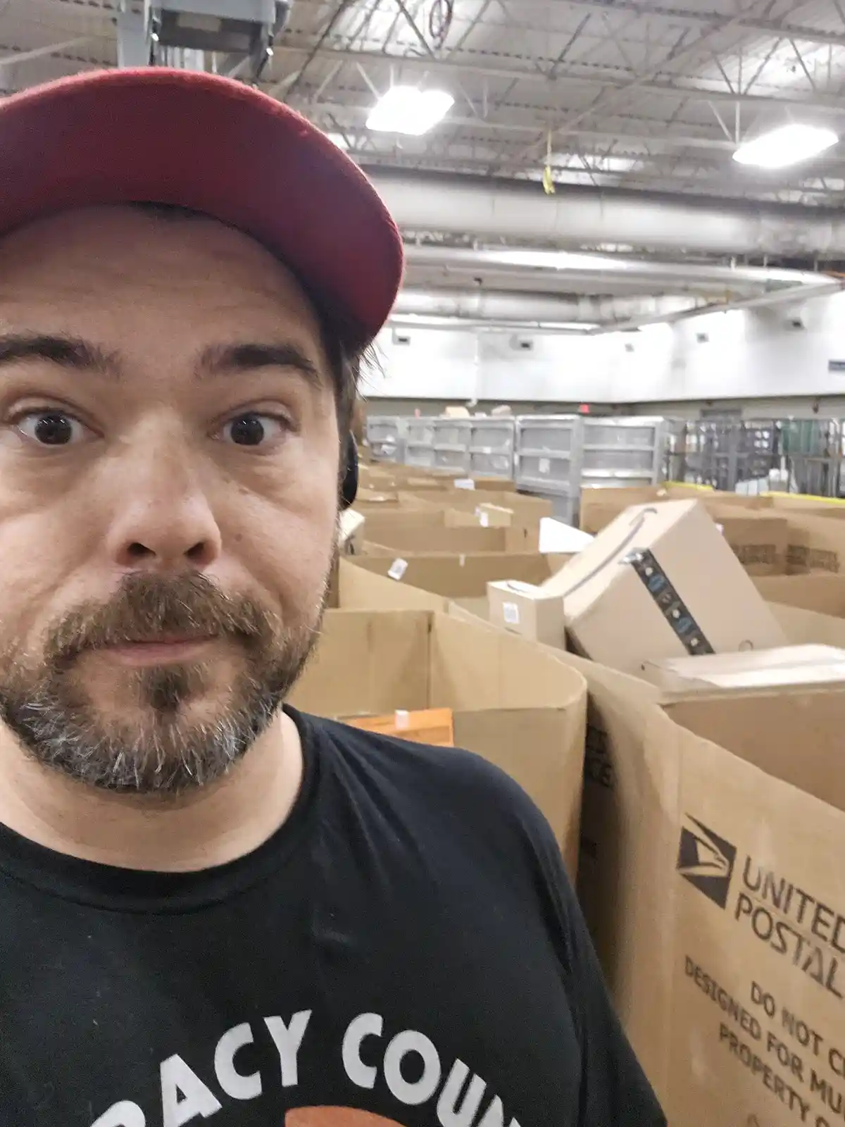 A man with a red cap and beard takes a selfie in a warehouse filled with large United Postal Service cardboard boxes. He has…