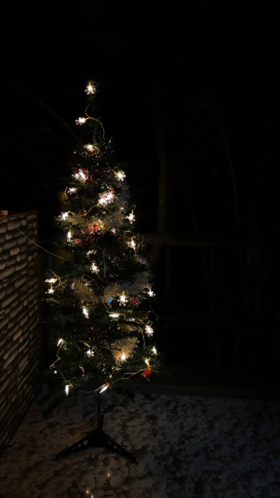 A small, decorated christmas tree with lights and ornaments stands alone on a pebble-covered floor at night, illuminated…