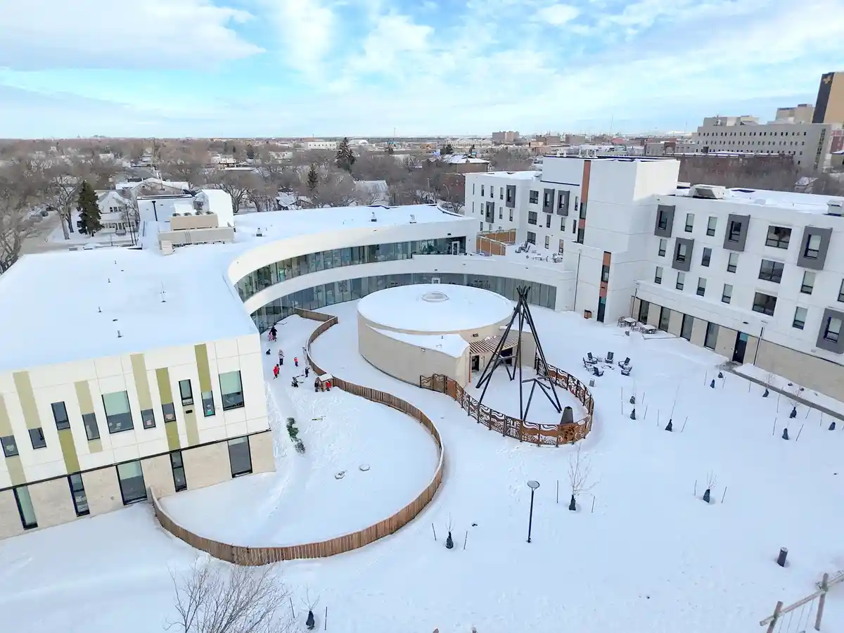 an aerial photo of a modern, snow-covered campus with curved buildings and a central courtyard. Several children are playing…