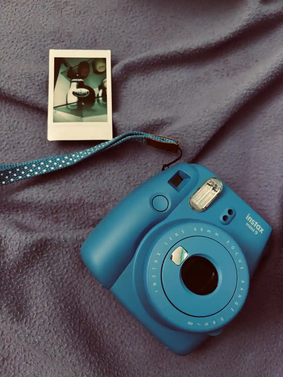A blue Fujifilm Instax Mini 9 instant camera with a blue and white polka-dot wrist strap rests on a textured purple blanket…
