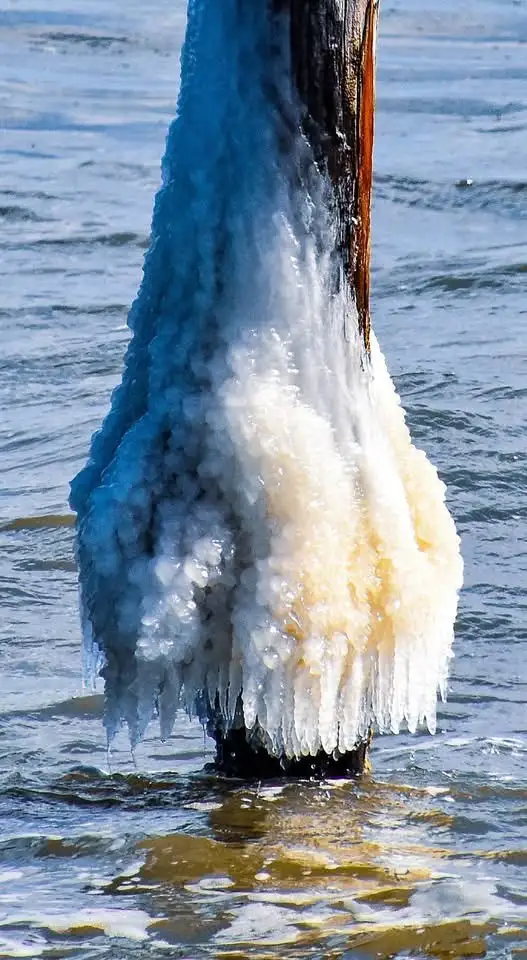 A vertical wooden post partially submerged in water, heavily encrusted with ice. The ice forms two distinct sections: a…