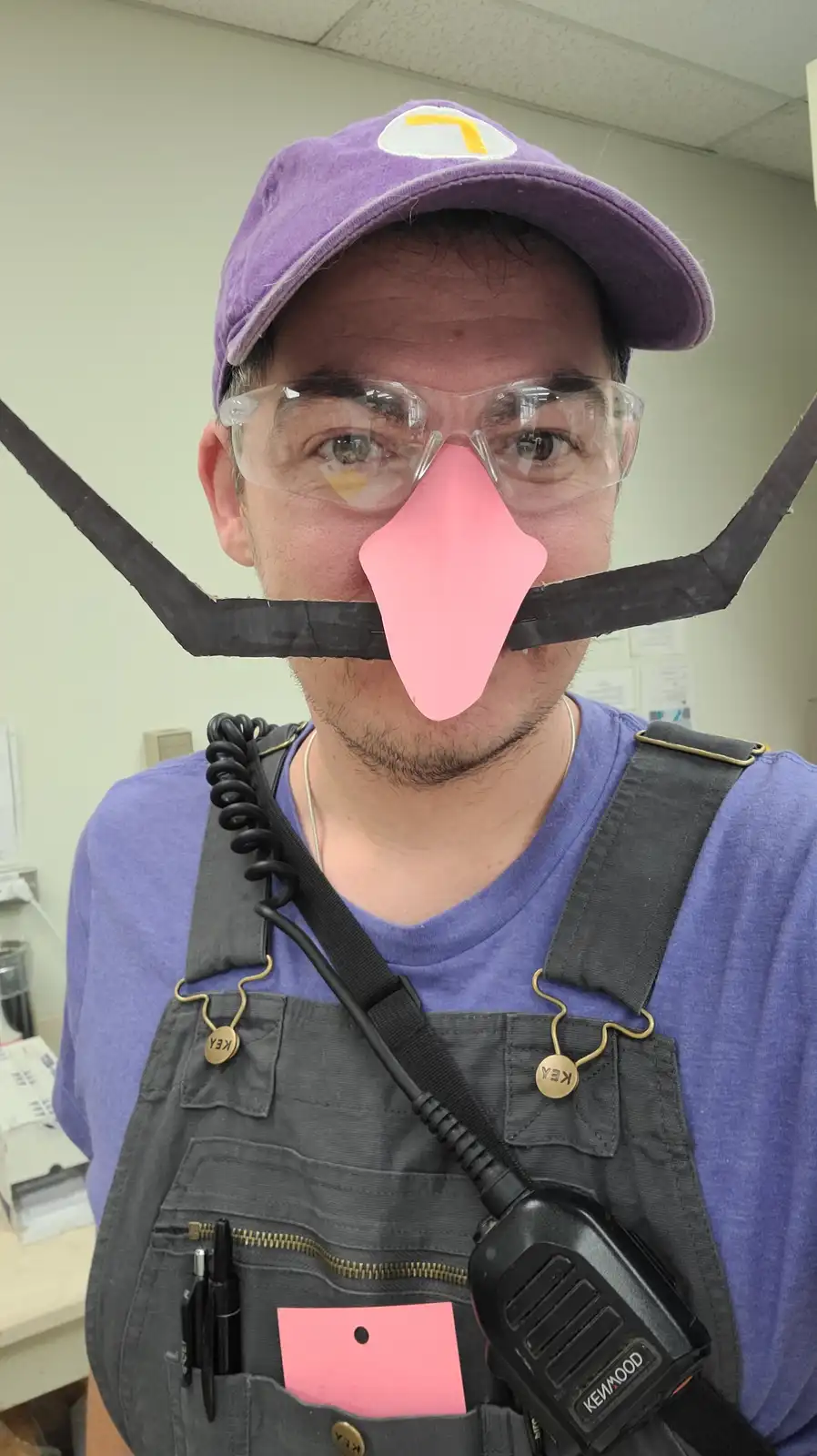 A man in a purple cap with a yellow 'W' logo, safety glasses, and grey overalls with a Kenwood radio, has a pink paper nose…