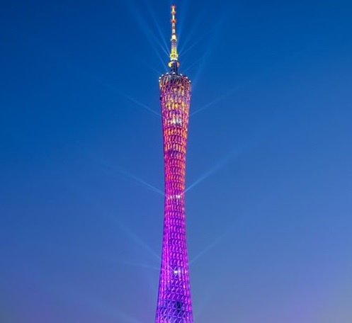 A tall, slender tower with a lattice-like structure is illuminated with vibrant purple and orange lights against a clear…