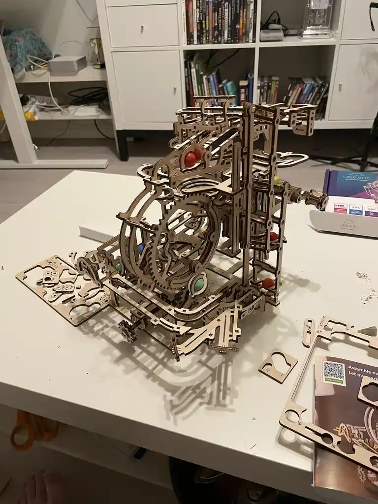 A complex wooden mechanical model, likely a UGEARS kit, sits partially assembled on a white table with scattered laser-cut…