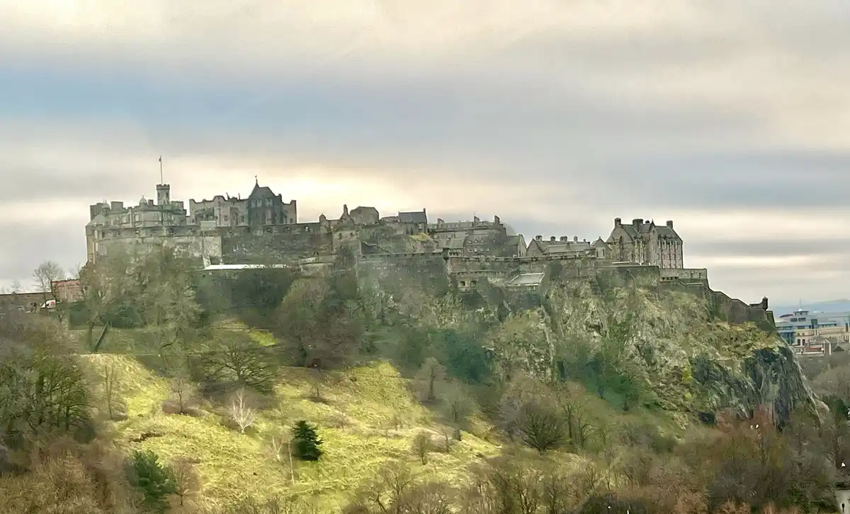 A wide-angle photograph shows Edinburgh Castle perched atop a steep, grassy, rocky hill under a cloudy sky. The castle's…