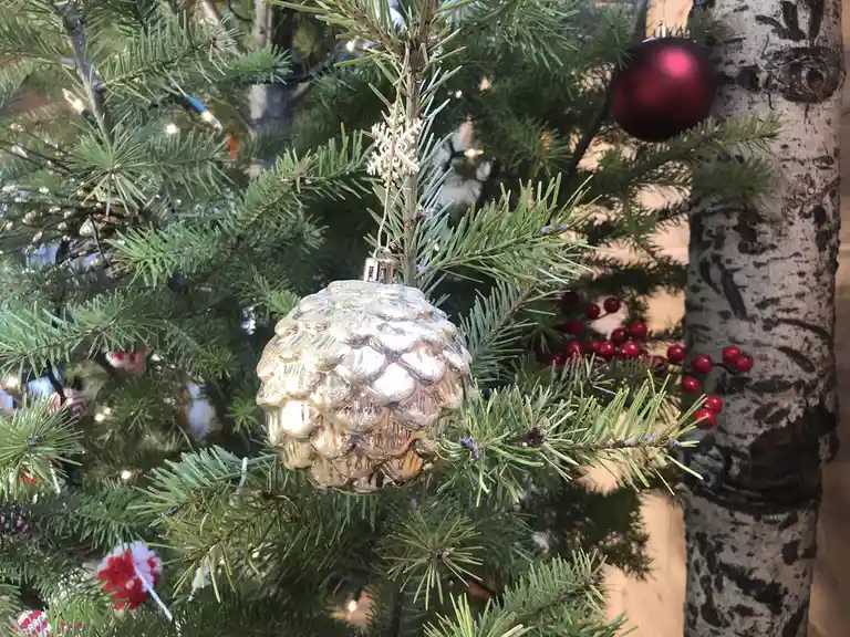 A close-up photo of a decorated evergreen tree shows a shiny, gold-colored pinecone-shaped ornament hanging from a branch…