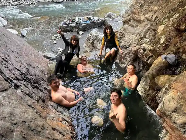 six people are posing cheerfully in a natural hot spring surrounded by rocks, with some sitting on the edge and others…