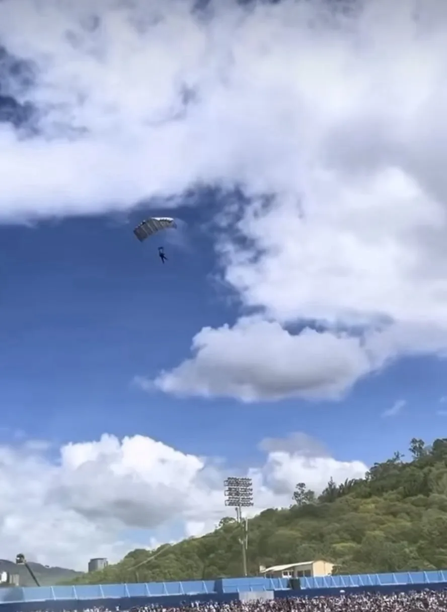 A parachutist with a large, patterned parachute descends slowly through a partly cloudy blue sky above a crowded stadium…