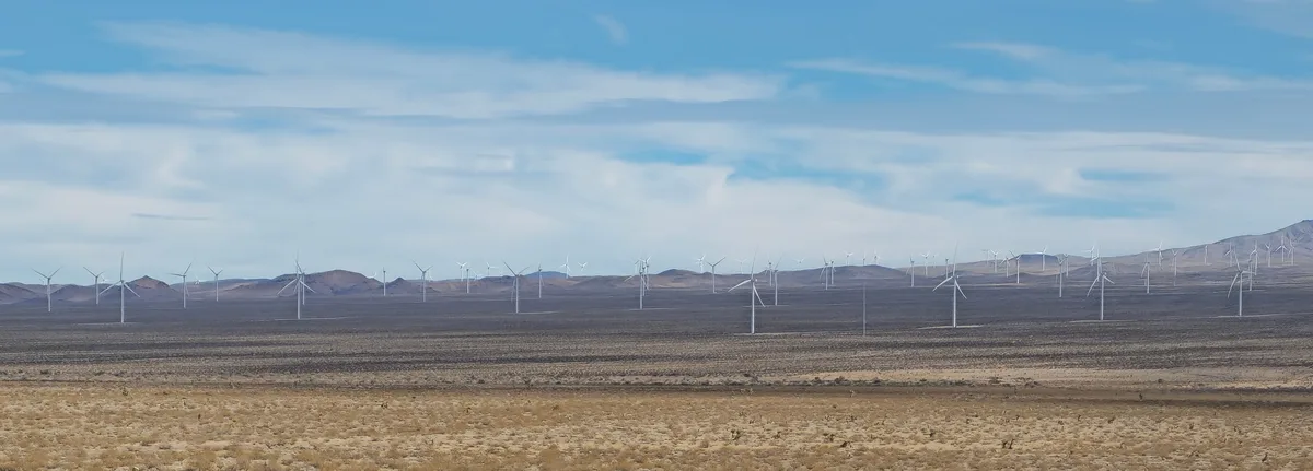 A wide landscape photo shows a vast desert with numerous white wind turbines stretching across rolling hills under a blue…