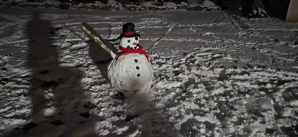 A snowman with a black top hat, red scarf, and twig arms stands in a snowy yard at night, with visible footprints and…