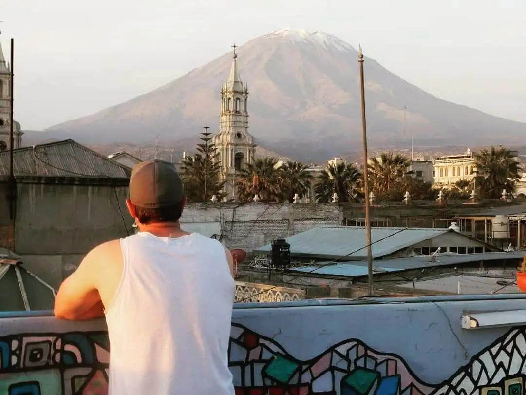A man in a white tank top and baseball cap stands on a rooftop, looking out at a cityscape with a large snow-capped mountain…