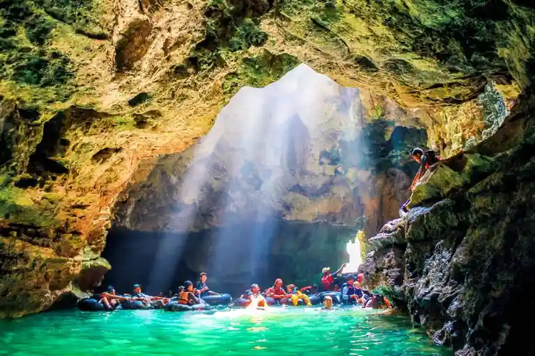 A group of people in inflatable tubes float in a vibrant turquoise cave pool, illuminated by dramatic beams of sunlight…