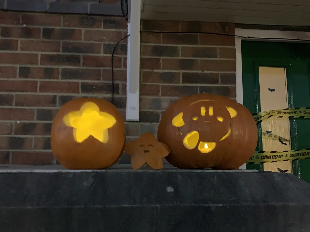 Two carved pumpkins sit on a porch step: one large pumpkin with a smiling face resembling Kirby, and a smaller pumpkin with…