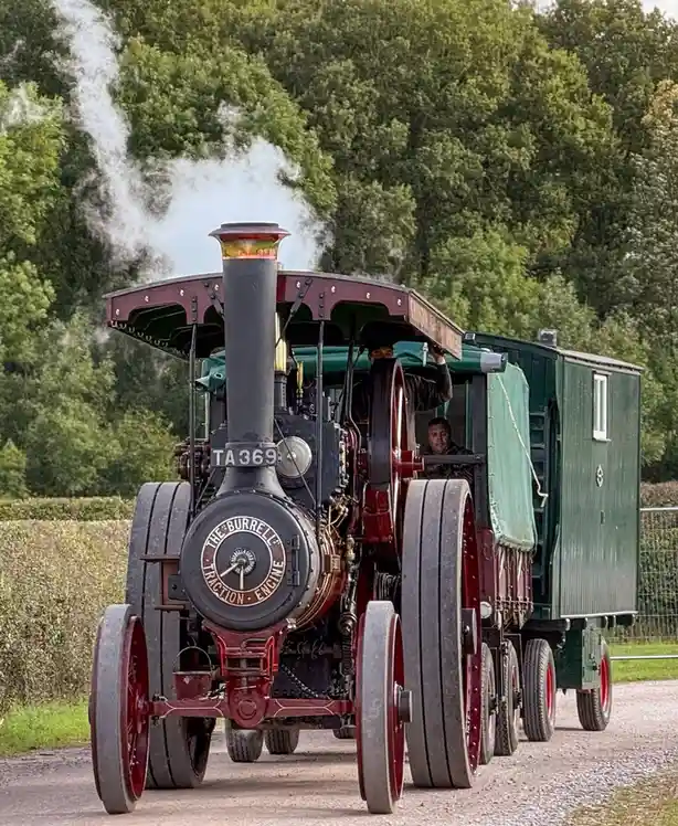 A vintage red and black steam traction engine, labeled 'THE BURRELL TRACTION ENGINE' on its front, emits white steam as it…