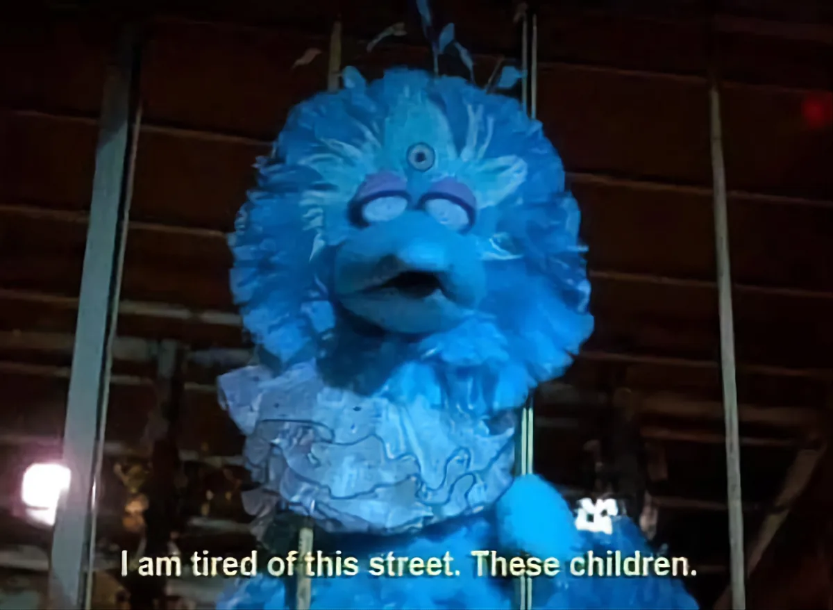 A blue, furry, three-eyed puppet with a weary expression is shown against a dark wooden background. The overlaid text reads…