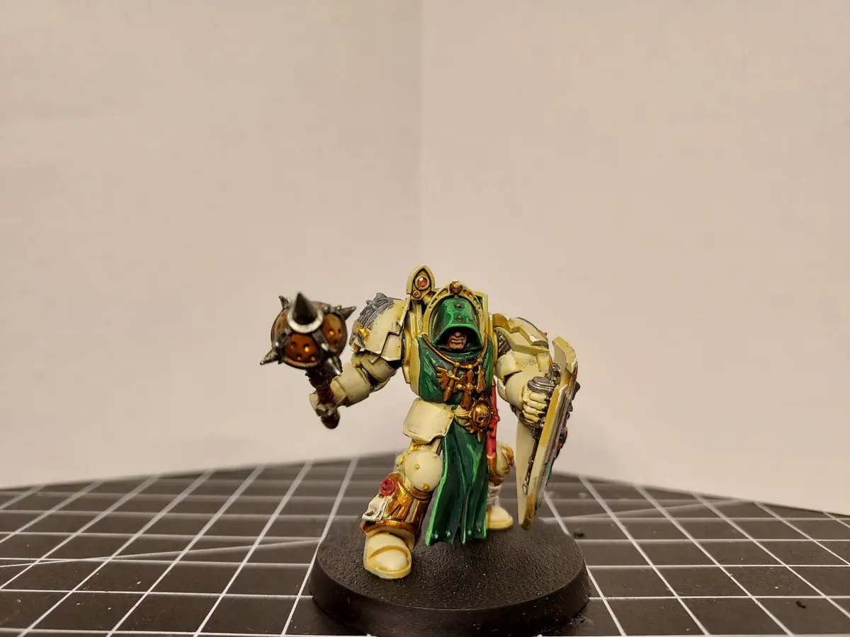 A painted miniature of a Space Marine in green and cream armor, holding a spiked mace and shield, stands on a black circular…