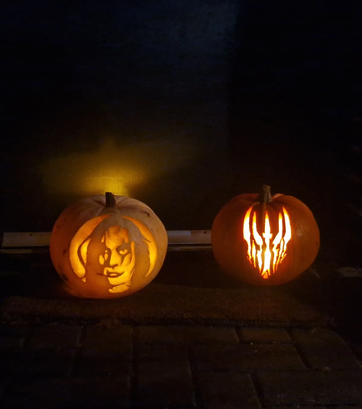 Two carved pumpkins sit side by side on a dark surface, illuminated from within. The left pumpkin features a detailed…