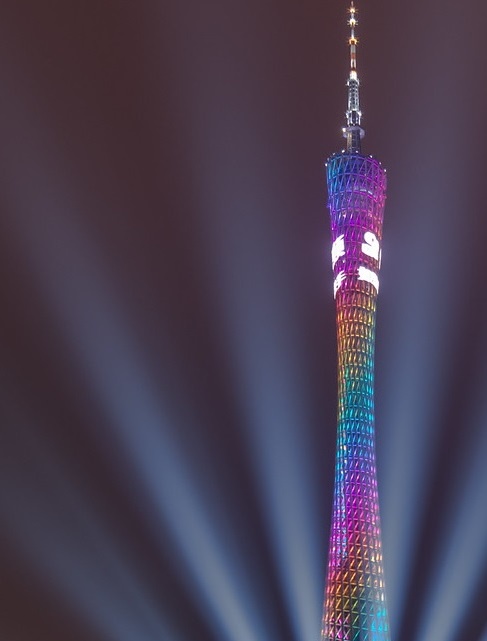 A tall, slender tower with a lattice-like structure is illuminated with a rainbow of colors at night, with beams of light…