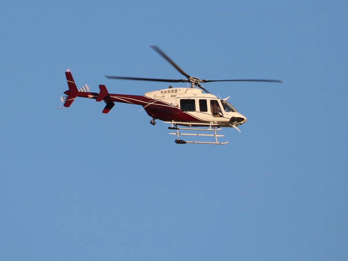 A white and maroon helicopter with registration N407BB flies against a clear blue sky, its rotors spinning. The image has…