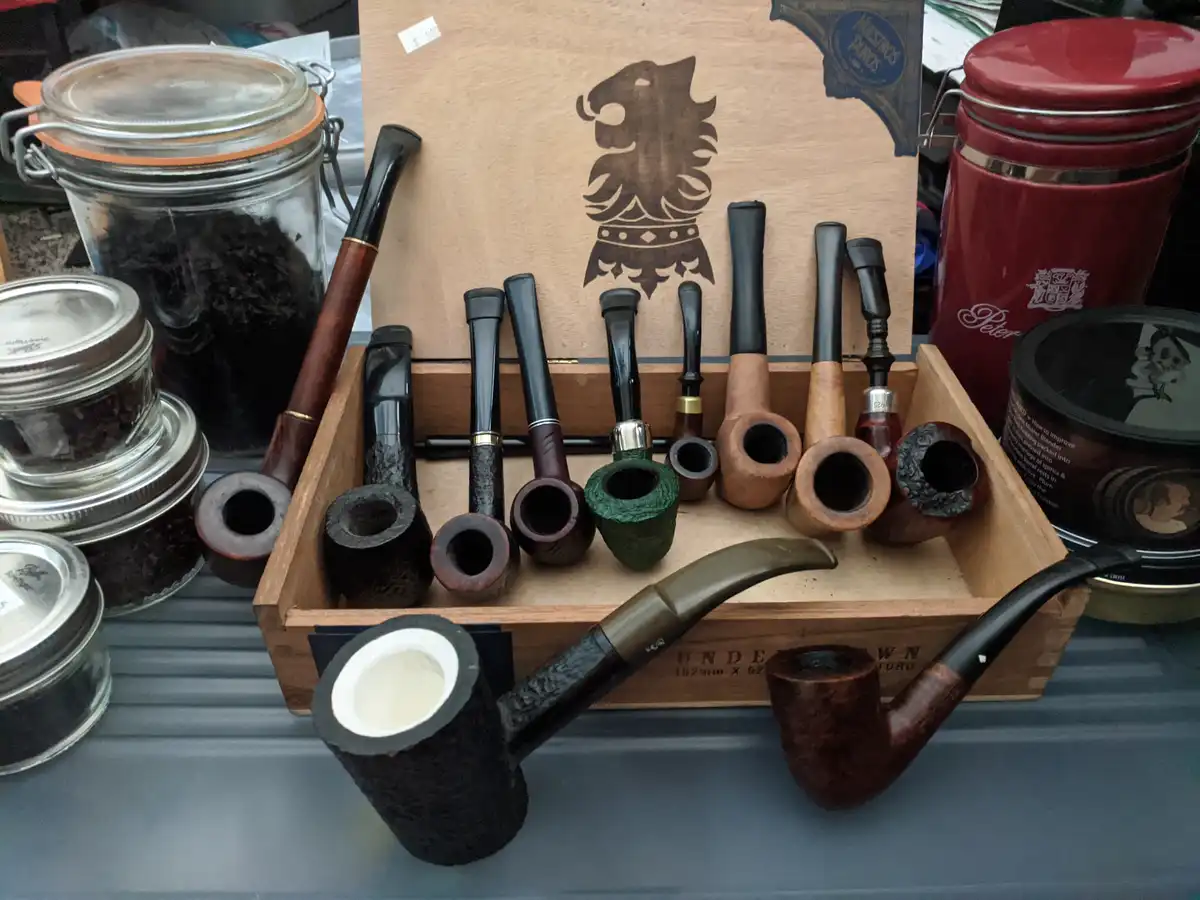 A wooden box filled with various smoking pipes of different shapes, sizes, and materials, including briar and clay…