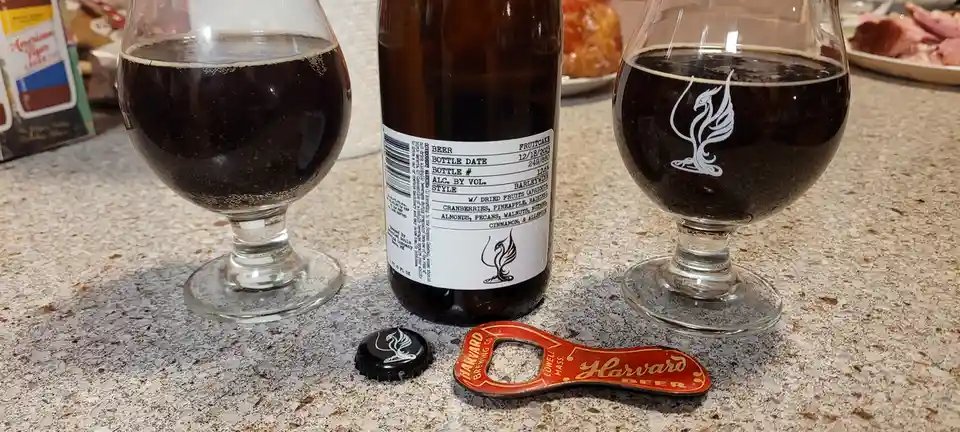 A bottle of 'Fruitcake' barleywine beer sits between two glasses filled with dark beer on a speckled countertop, with a…