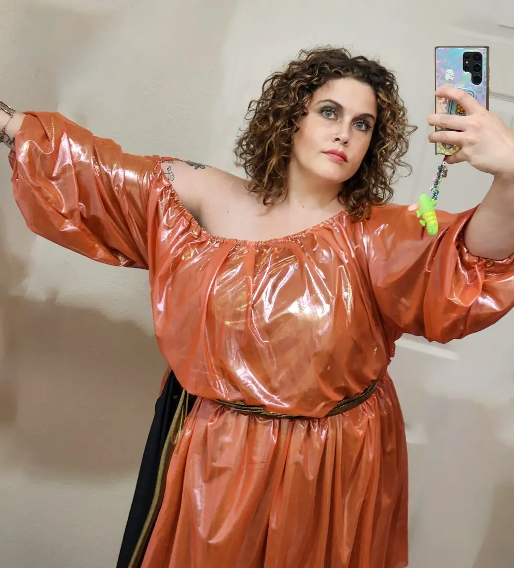 A woman with curly hair takes a mirror selfie wearing a shiny, orange, off-the-shoulder dress with puffy sleeves and a black…
