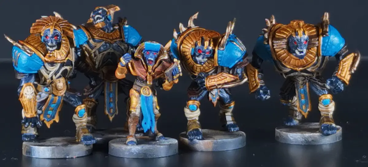 Five painted miniature figurines of blue-skinned, red-eyed, Egyptian-themed warriors in gold and blue armor stand on…
