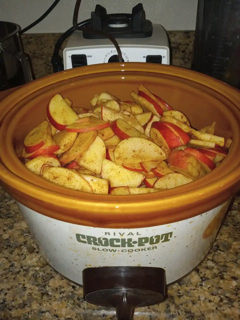 A Rival Crock-Pot slow cooker is filled with sliced red apples coated in spices, sitting on a granite countertop with a…
