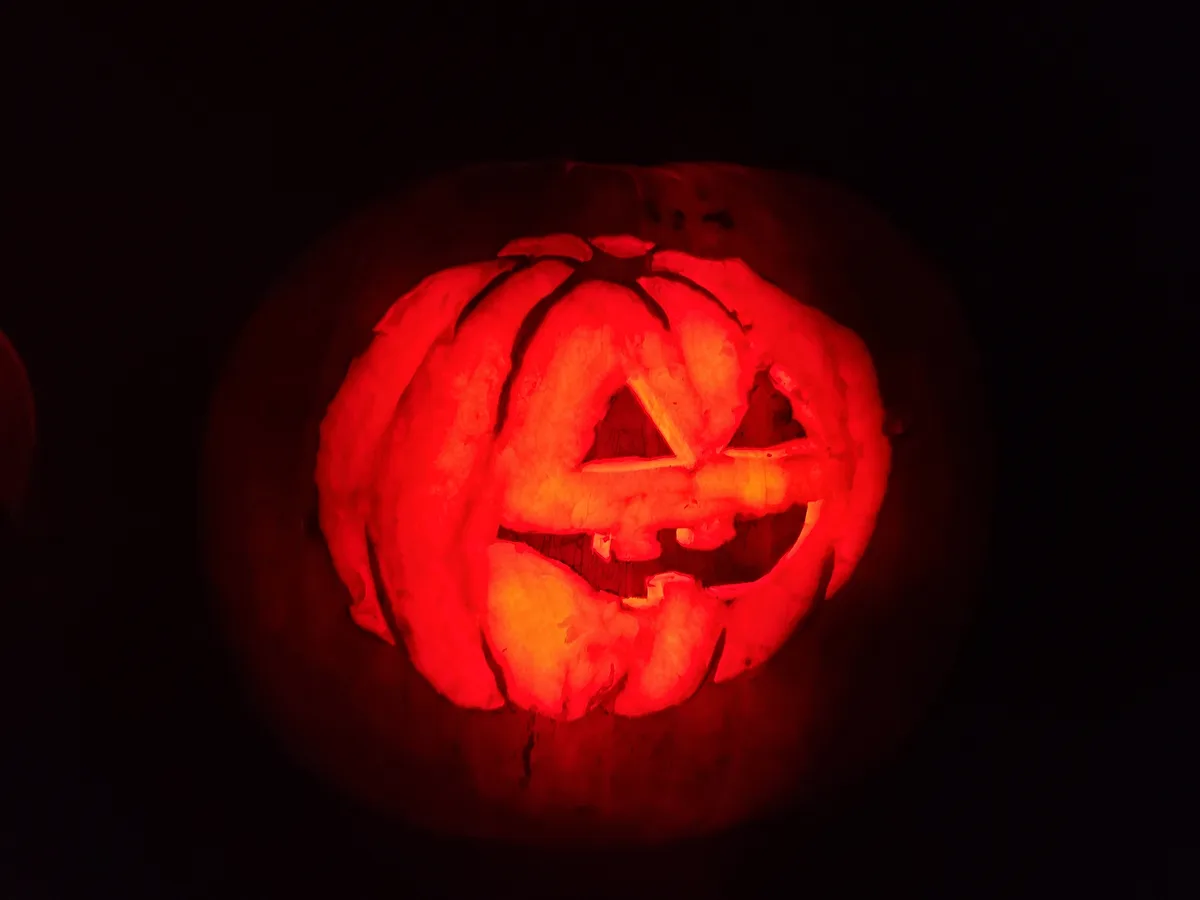 A carved pumpkin, illuminated from within by a red light, displays a face with triangular eyes and a jagged mouth. The…
