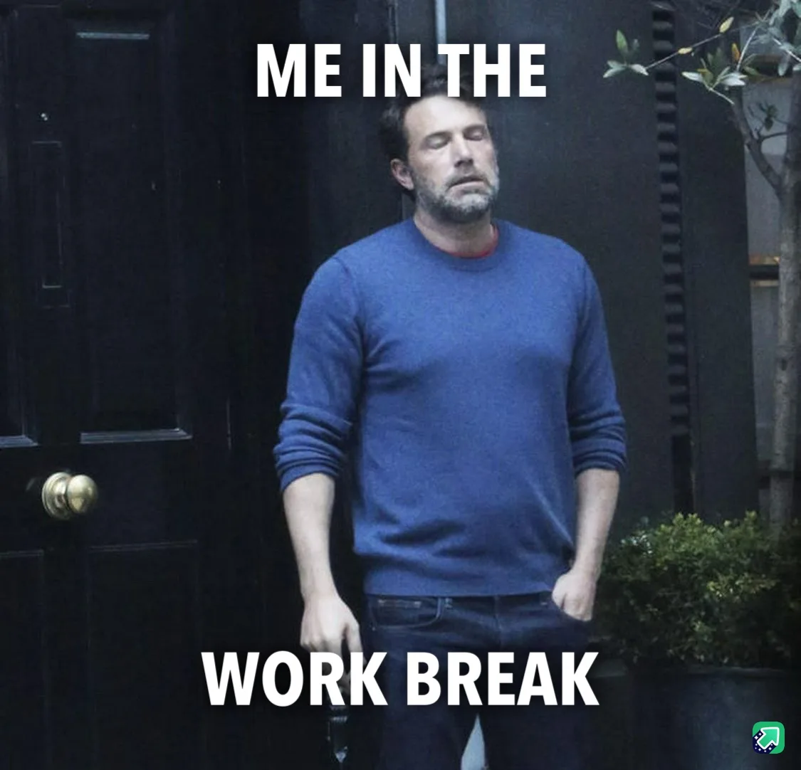 A man with a beard and closed eyes stands in front of a dark door, wearing a blue sweater and jeans, looking exhausted. The…