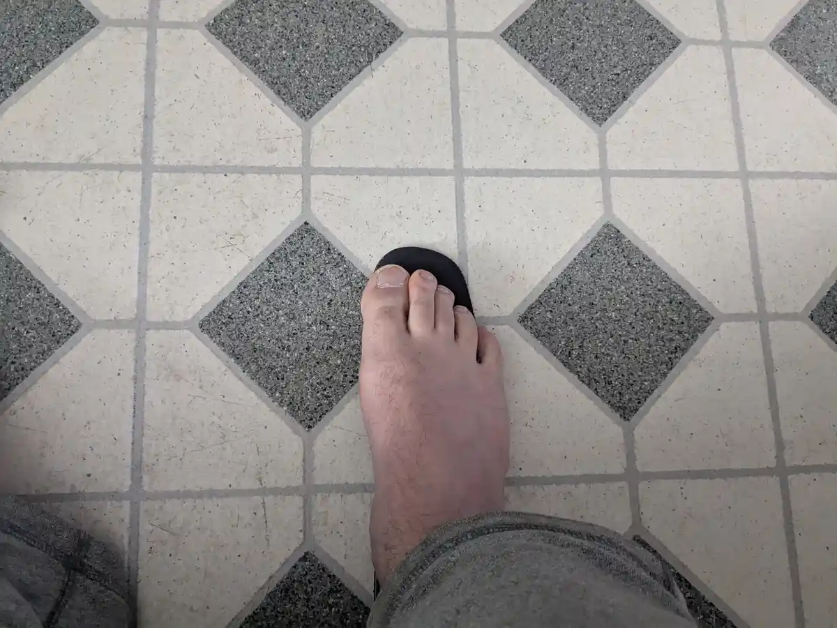 A top-down view of a person's bare foot wearing a black flip flop, standing on a tiled floor with a repeating pattern of…