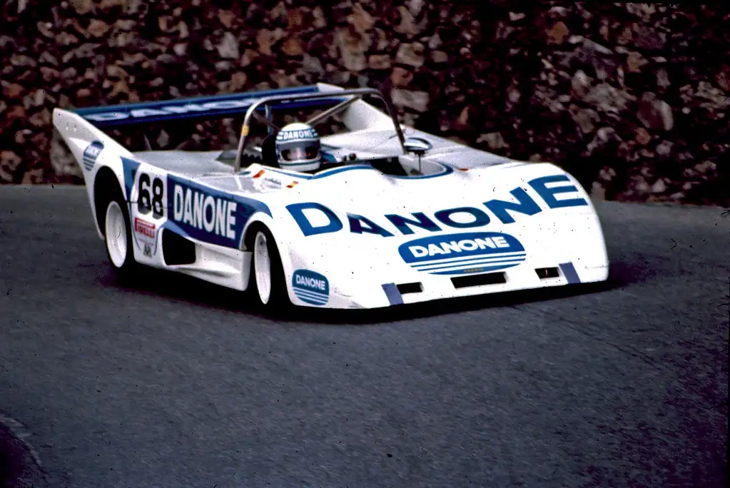 A vintage white and blue Danone-sponsored race car, number 68, speeds along a track with a rocky wall in the background. The…