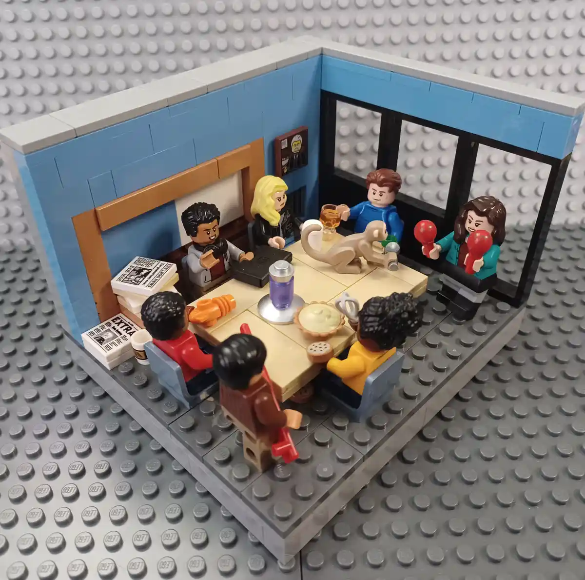 A LEGO diorama depicts a living room scene with multiple LEGO minifigures seated around a table, resembling characters from…