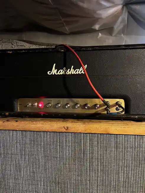 A close-up photo of a black Marshall guitar amplifier head with a gold control panel. A red power cable is plugged into the…