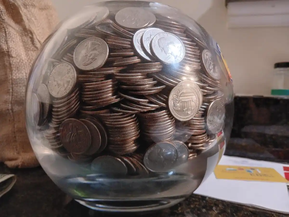 A clear glass globe filled with stacks of U.S. coins, mostly quarters and dimes, sits on a countertop. The overlaid text…