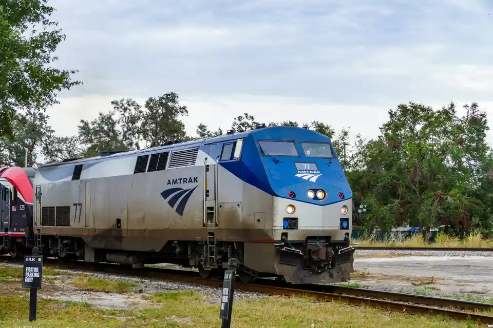 An Amtrak train engine, numbered 77, is pictured on railway tracks with trees in the background. In the foreground, a black…