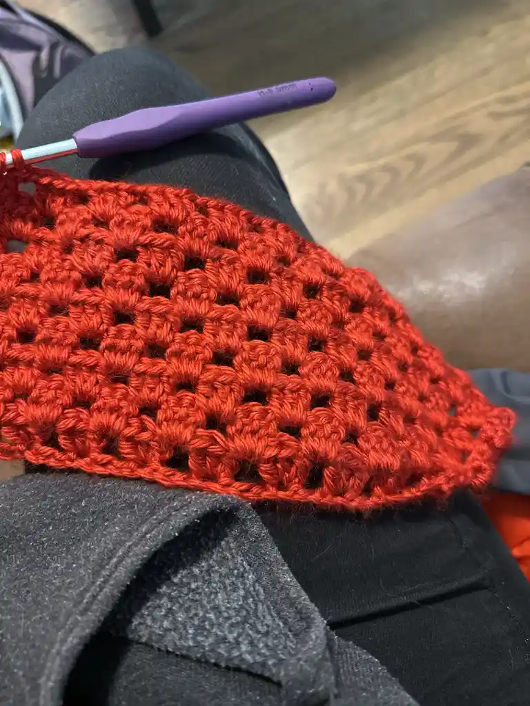 A close-up photo shows a bright red crocheted fabric with a textured pattern resting on someone's lap, which is covered in…