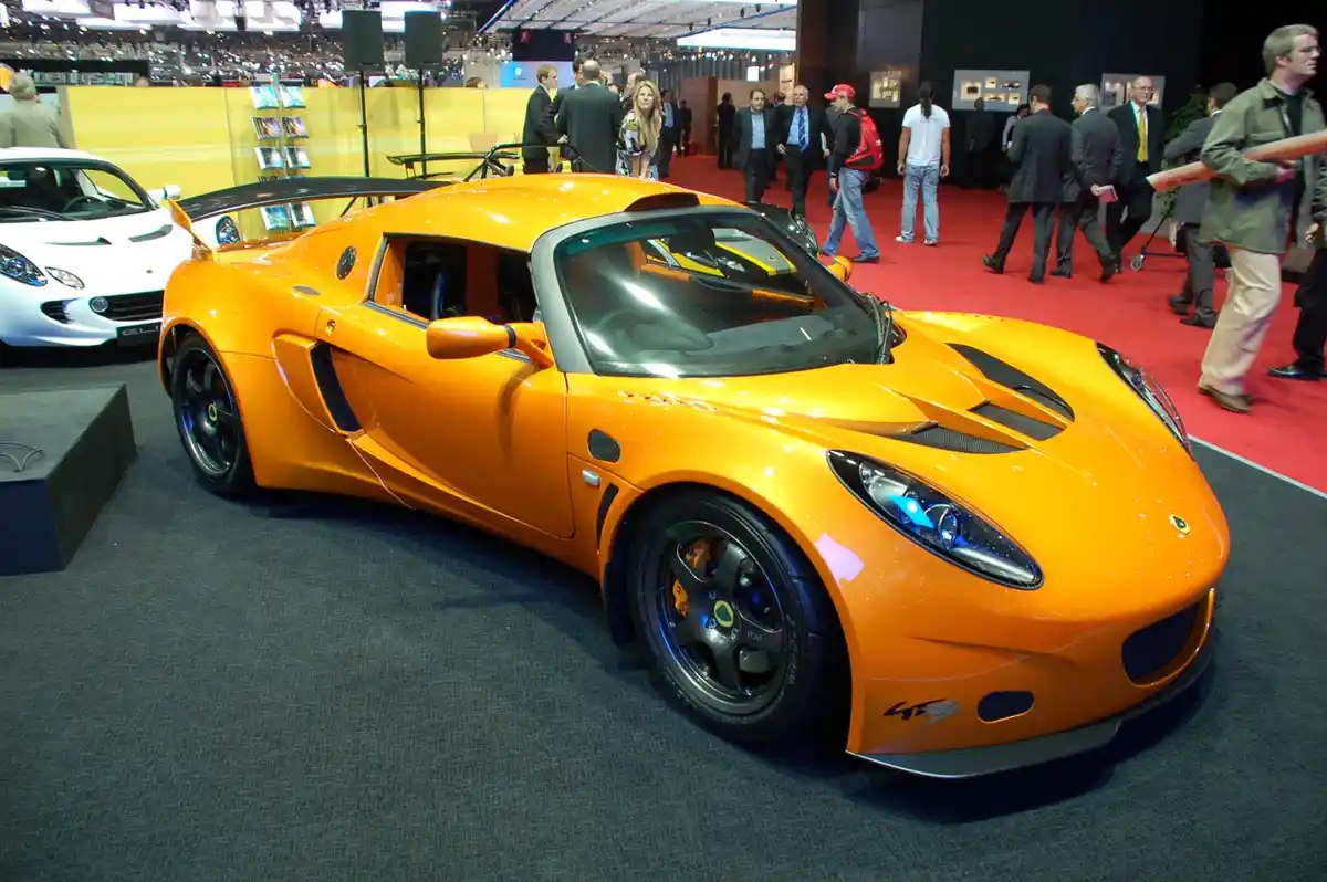 a bright orange lotus exige gt4 sports car is displayed at an auto show on a dark gray carpet, with people walking around on…