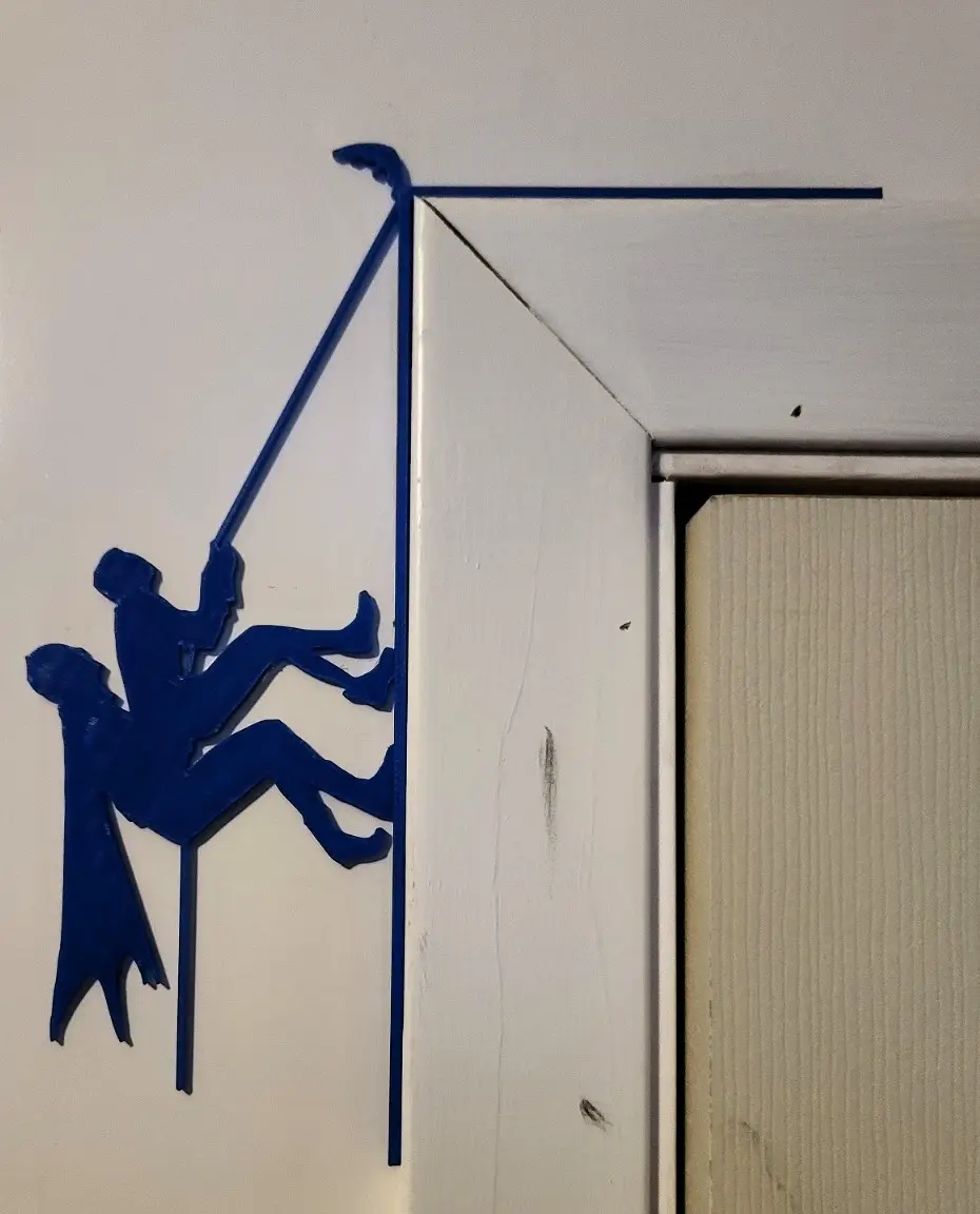 A blue 3D-printed silhouette of two people climbing a rope is mounted on a white wall next to a door frame. The figure at…