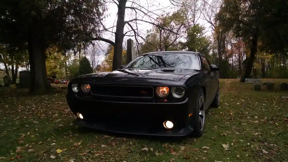 A black Dodge Challenger RT is parked on grass in a cemetery, surrounded by tombstones and autumn trees. The car's…