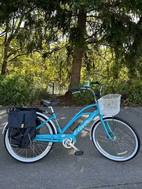 A bright turquoise Electra bicycle with white fenders, a white wire basket on the front, and a black pannier bag on the rear…