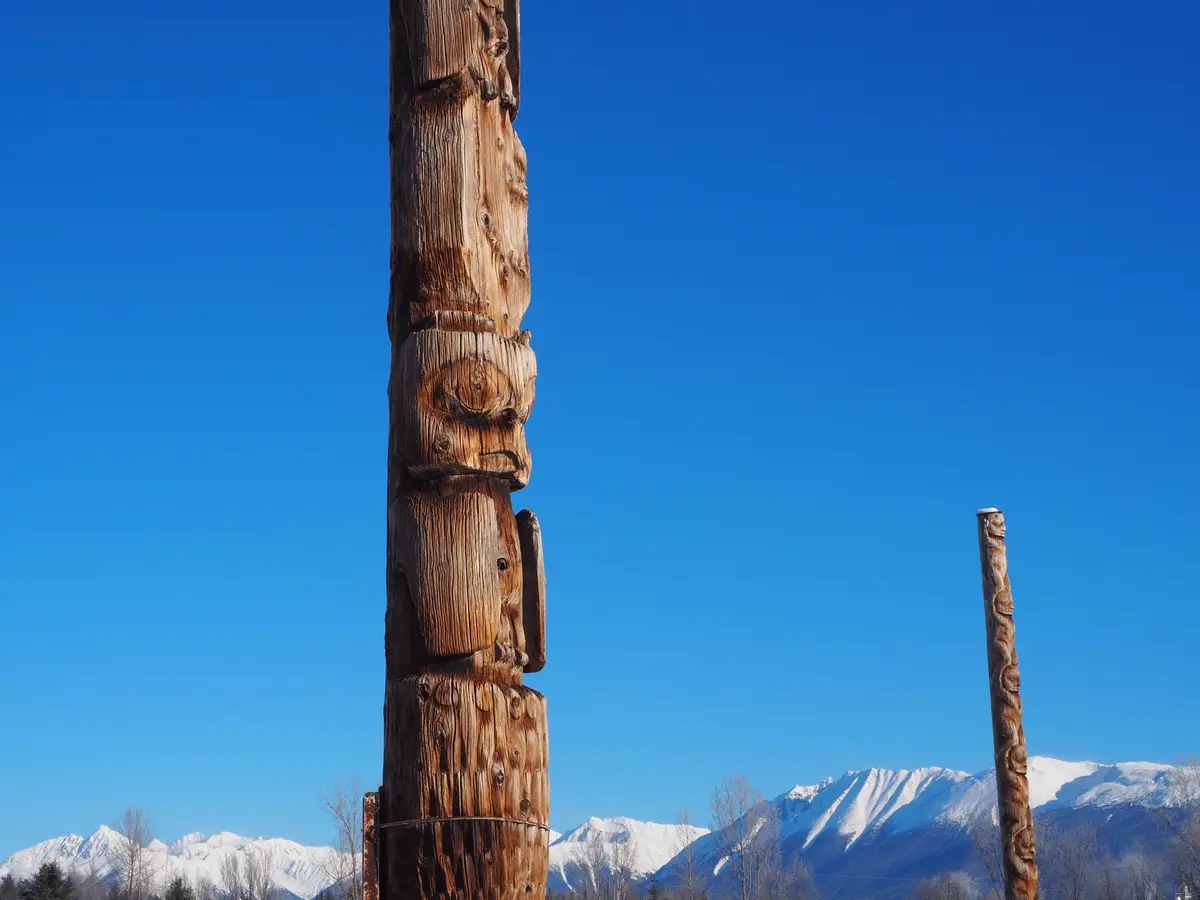 A tall, intricately carved wooden totem pole stands prominently against a clear blue sky, with snow-capped mountains visible…