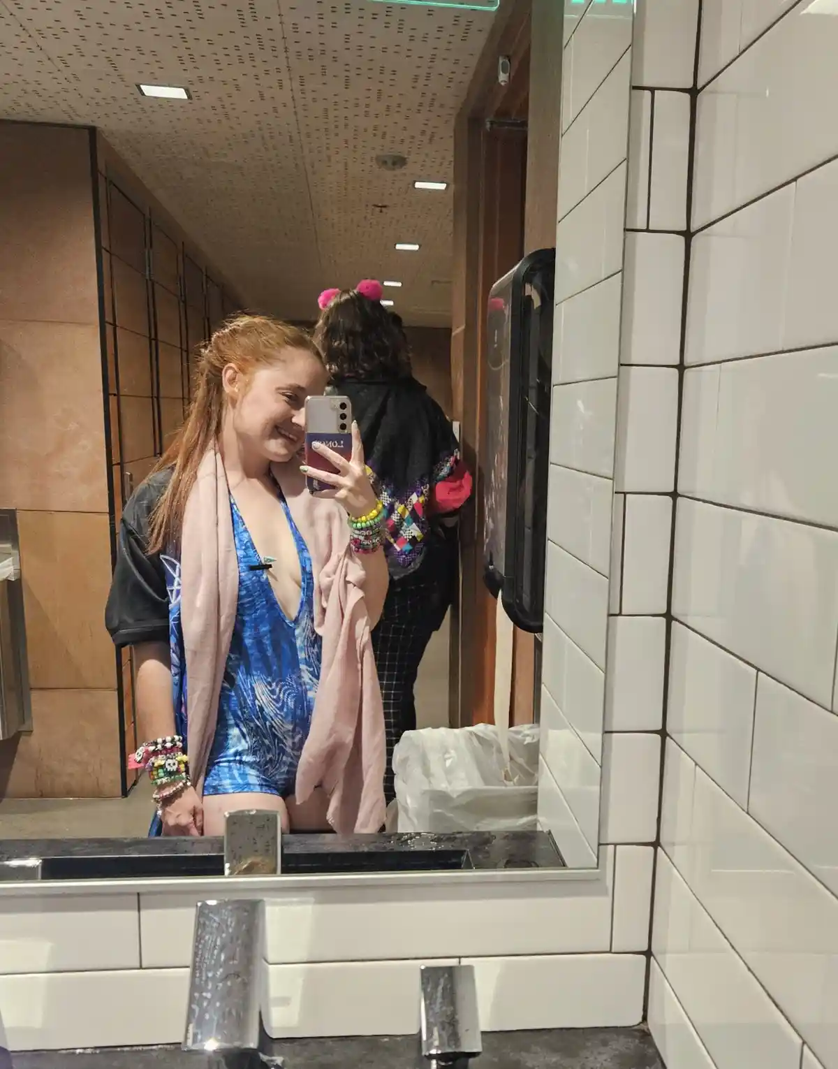 A young woman with red hair, wearing a blue tie-dye swimsuit and a pink towel draped over her shoulders, takes a selfie in a…