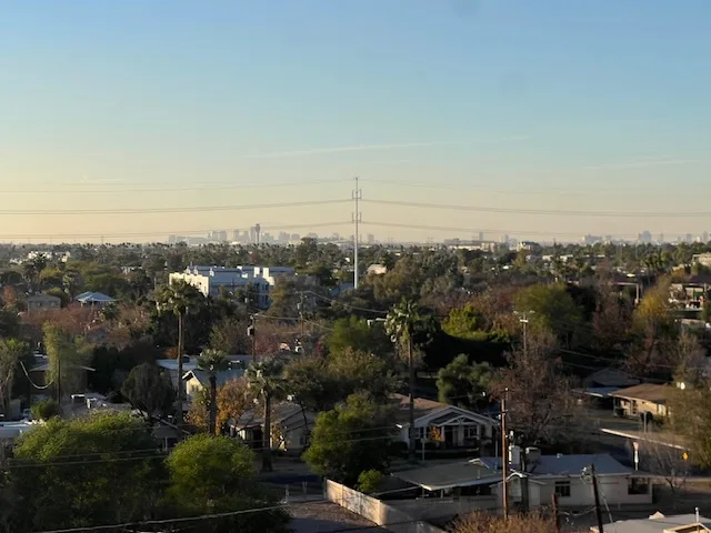 A daytime photo of a suburban neighborhood with houses, trees, and power lines, with a distant city skyline visible on the…