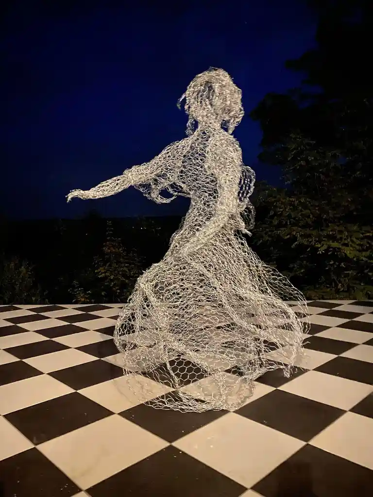 A sculpture of a dancing woman made entirely of chicken wire stands on a black-and-white checkered floor at night, with…