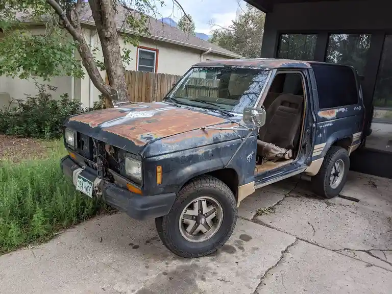 A heavily rusted, old blue SUV with its driver's door open and interior seats exposed sits on a cracked concrete driveway…