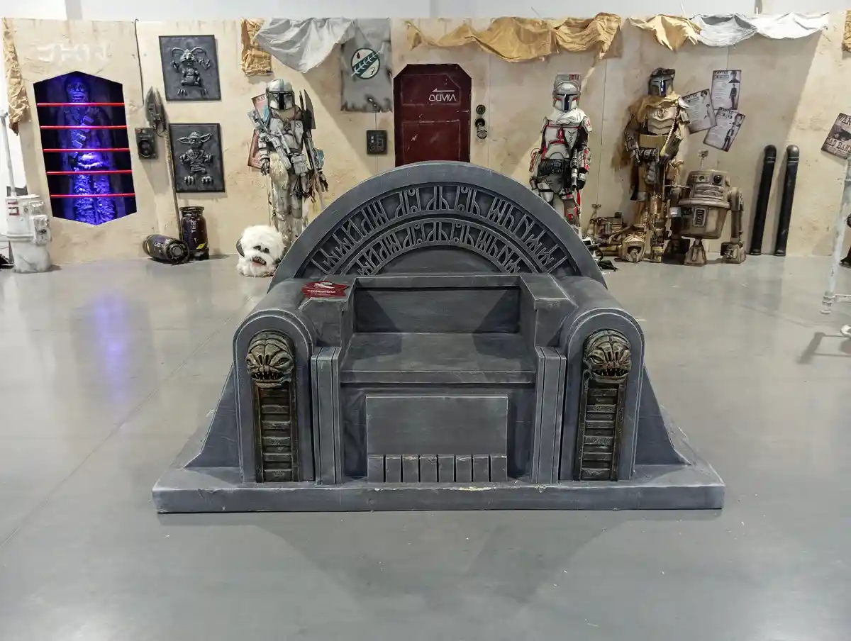 a large, empty, dark gray throne with ornate carvings and snake-head motifs on its armrests sits center stage in a star…