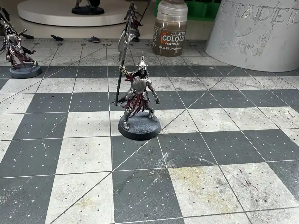 A painted miniature of a skeletal knight in silver armor holding a large axe stands on a checkered gaming mat, surrounded by…