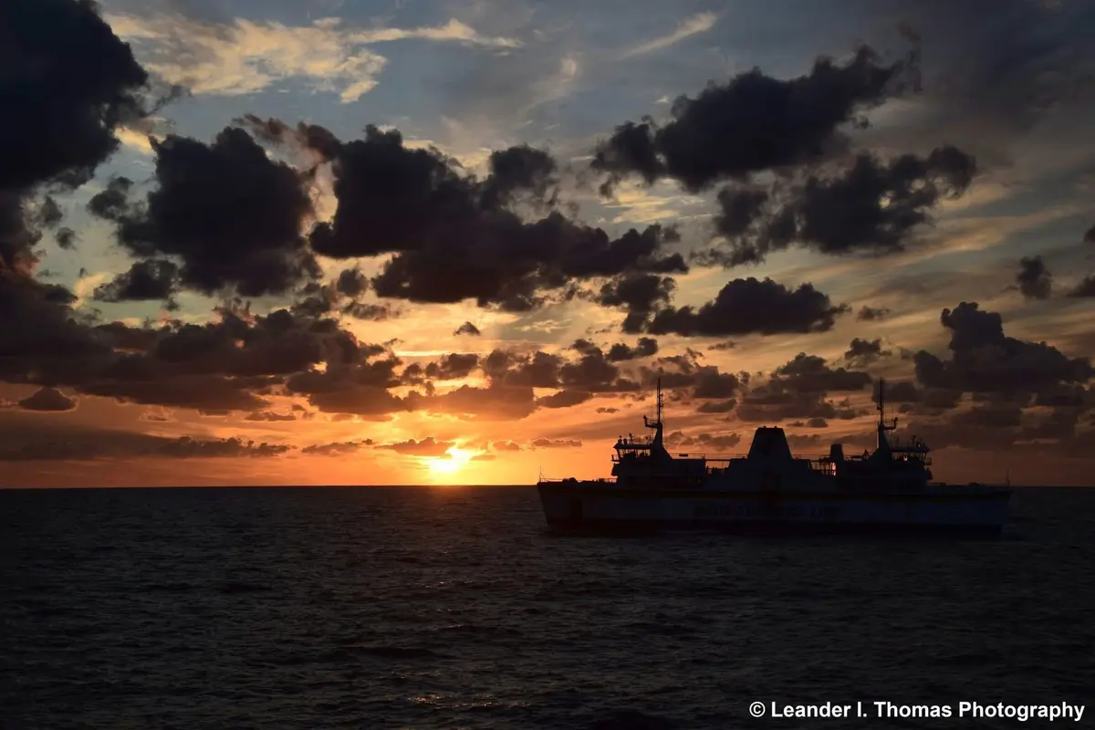 A silhouette of a ferry labeled 'GOZO CHANNEL LINE' sails on the ocean during a dramatic sunset, with the sun low on the…